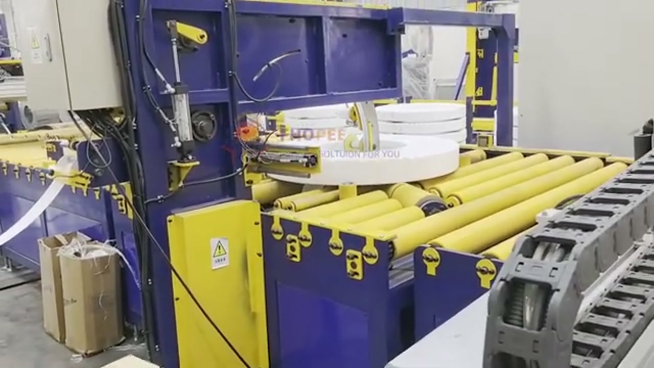 Film-wrapped steel coil lying flat on yellow roller conveyors, seen in a single-camera view