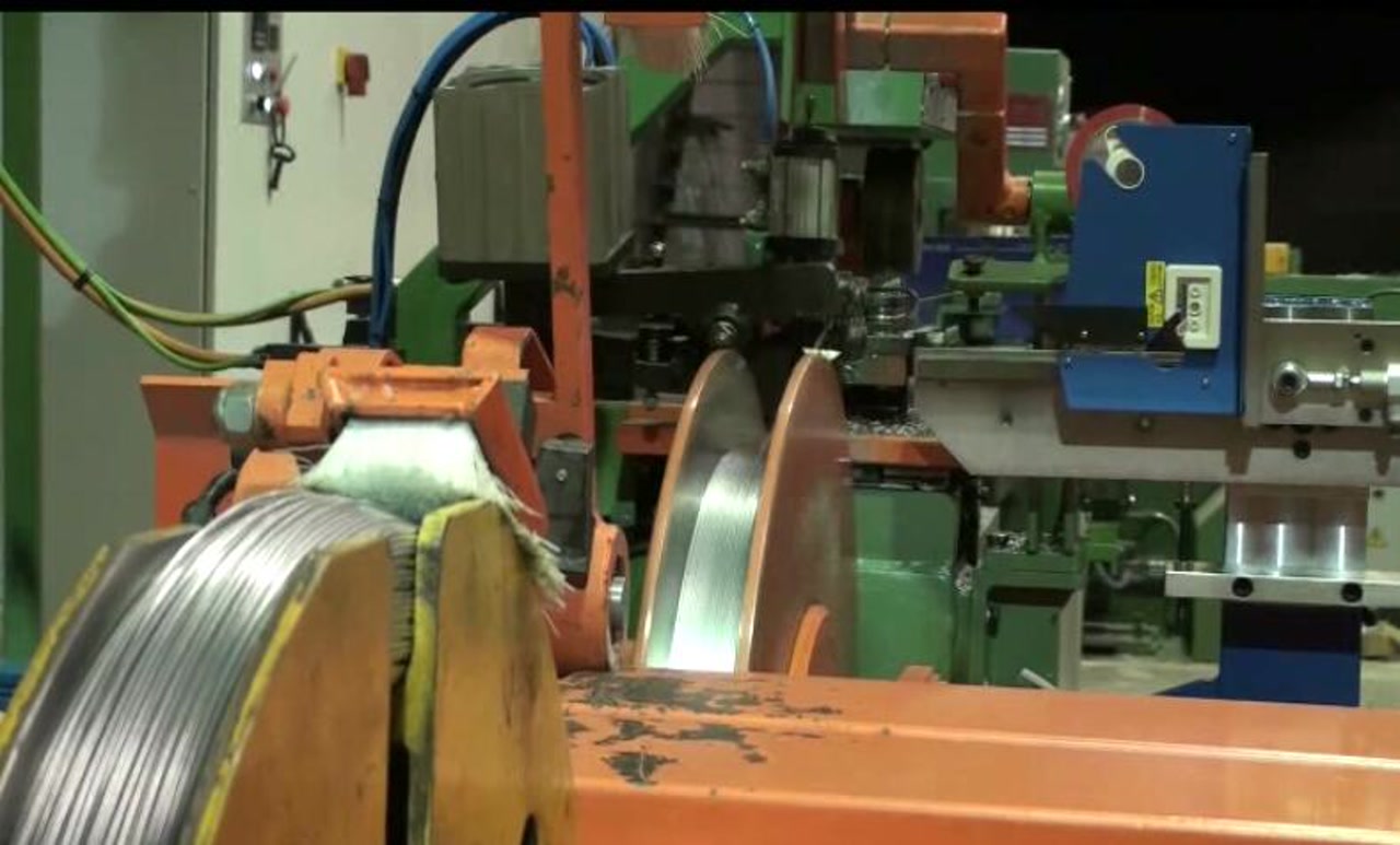 A wire coiling and winding machine with a feed coil on an orange cradle and large flanged winding discs